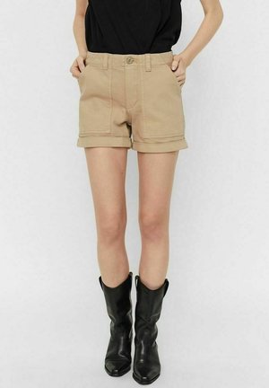 Mujer con pantalones cortos beige con puños y bolsillos delanteros y botas negras de cuero hasta la mitad de la pantorrilla, de pie con las manos en los bolsillos.