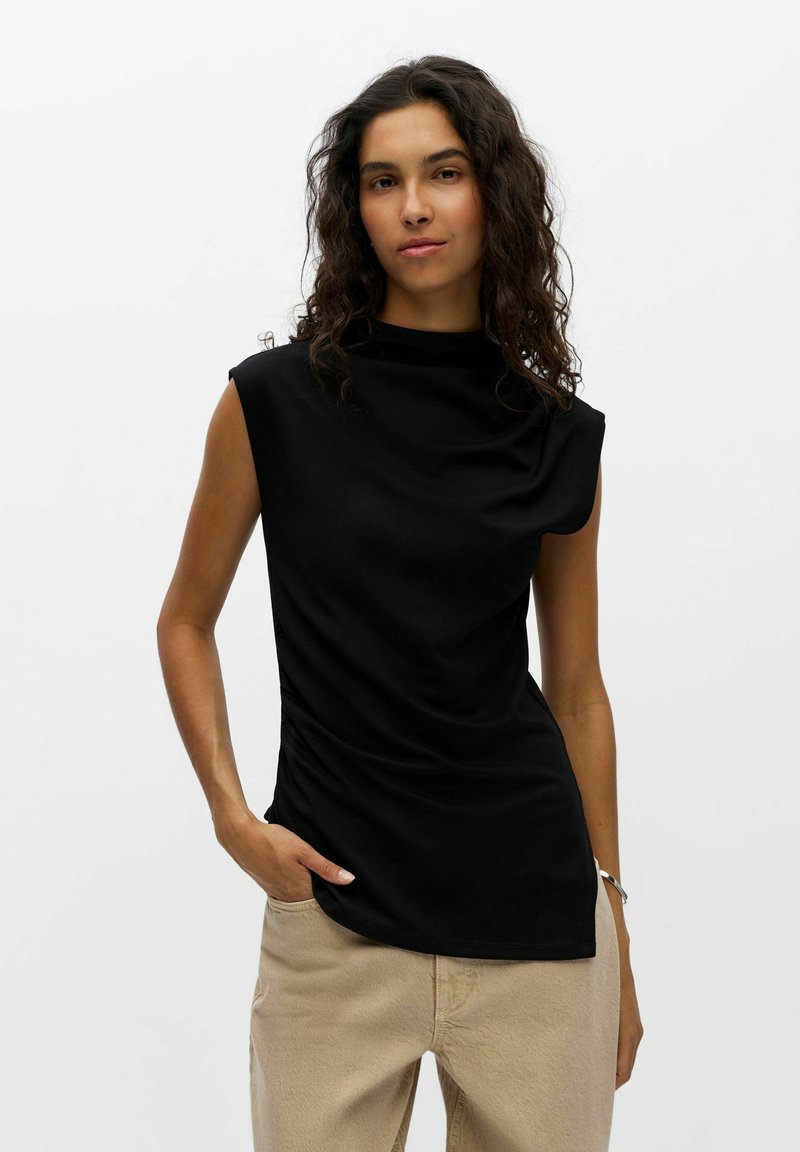 Femme aux cheveux bouclés portant un haut noir sans manches et un pantalon beige, debout avec une main dans la poche sur un fond uni.