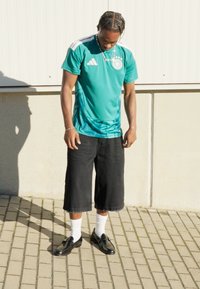 Een turquoise Adidas-shirt met witte accenten, gecombineerd met zwarte knielange shorts en zwarte schoenen op witte sokken, op een verharde ondergrond.