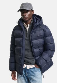 Marineblå puffjakke med avtakbar hette, quiltet design og glidelås. Brukt over en grå genser og rutete skjorte, sammen med lyseblå jeans.