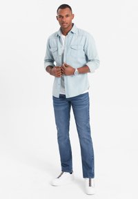 Camisa de mezclilla azul claro de botones superpuesta sobre una camiseta blanca, combinada con jeans azul oscuro y zapatillas blancas con detalles a rayas.