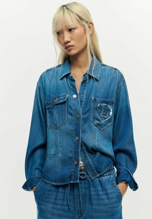 Young woman with long blonde hair wearing a blue denim jacket and matching pants, standing with hands in pockets against a plain background.