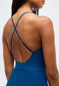 Γυναίκα με πλεκτά μαλλιά που φοράει αμάνικο μπλε φόρεμα με λεπτούς, ρυθμιζόμενους σταυρωτούς ιμάντες και χαμηλή πλάτη.