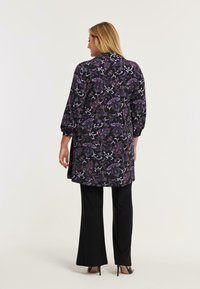 Kvinde set bagfra i en langærmet mørk tunika med lilla paisley-mønster, sorte brede bukser og sorte højhælede sko foran en ensfarvet baggrund.