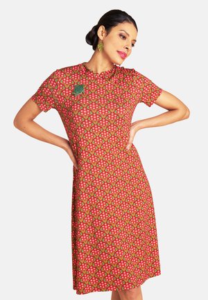 Frau mit dunkel gebundenem Haar, die ein rotes, kurzärmliges Kleid mit Blumenmuster, eine grüne Brosche und goldene Ohrringe trägt, steht mit den Händen in der Hüfte.