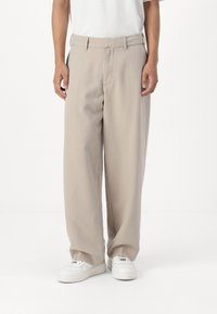 Pantaloni beige a gamba larga realizzati in tessuto liscio, con parte anteriore piatta, passanti per cintura e tasche laterali, abbinati a sneakers bianche.
