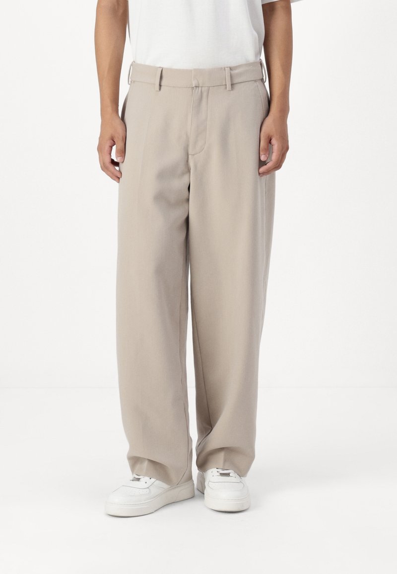 Pantaloni beige a gamba larga realizzati in tessuto liscio, con parte anteriore piatta, passanti per cintura e tasche laterali, abbinati a sneakers bianche.