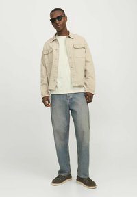 Jachetă din denim bej peste un tricou verde deschis, blugi albaștri și ochelari de soare negri. Ținută casual cu buzunare frontale și croială relaxată.