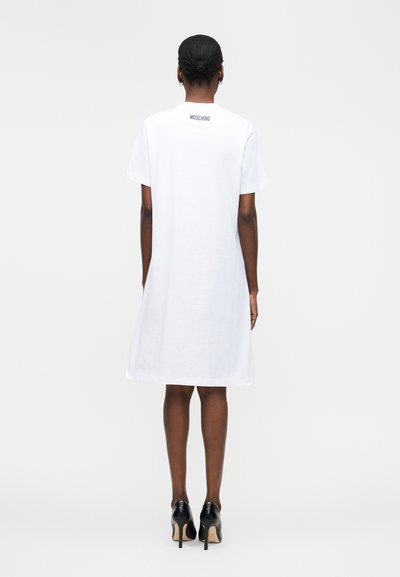 Femme à la peau foncée et aux cheveux attachés en chignon, portant une robe blanche Moschino mi-longue et des chaussures à talons noires, debout dos à la caméra.