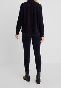 Persona che indossa una camicia e pantaloni di corduroy neri con stivaletti neri con tacco, in piedi di spalle su uno sfondo chiaro e semplice.