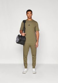 Αθλητική ένδυση σε ελαιόχρωμο με μαύρες πλαϊνές ρίγες, κοντομάνικο πουκάμισο και εφαρμοστά παντελόνια. Μαύρη και γκρι τσάντα με λεπτομέρειες από δίχτυ. Λευκά αθλητικά παπούτσια.