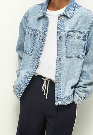 Veste en jean bleu clair avec poches avant portée sur un t-shirt blanc et un pantalon noir à cordon, mains dans les poches.