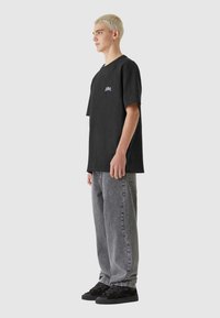 Črni oversize majica s majhnim vijoličastim grafičnim motivom na prsih, v kombinaciji s svetlo sivo baggy jeans hlačami in črnimi športnimi čevlji.