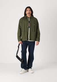 Veste légère vert olive avec fermeture à boutons et poche poitrine, portée sur une chemise blanche avec un logo, assortie à un pantalon marine et des baskets blanches.
