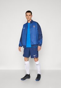 Giacca Nike blu con motivo a rombi, camicia blu, pantaloni corti blu scuro, calze bianche e scarpe da calcio nere. Presenta il logo PSG sulla giacca e sui pantaloni.