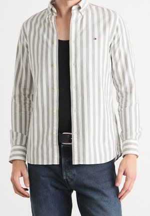 Mężczyzna w szaro-białej pionowo prążkowanej koszuli zapinanej na guziki, na czarnym topie i niebieskich dżinsach z czarnym paskiem, stojący na jednolitym tle.