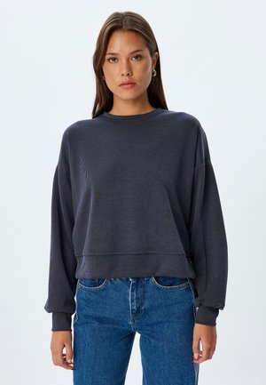 Sweatshirt grigio scuro a coste con un fit rilassato, scollatura rotonda e maniche lunghe. Abbinato a jeans in denim blu a vita alta.
