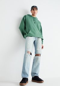 Grüne Kapuzenjacke mit Kängurutasche, hellblaue zerrissene Jeans und schwarze Sneakers mit Gummisohlen. Eine schlichte schwarze Mütze vervollständigt das Outfit.