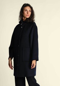 Cappotto in lana navy con colletto alto, chiusura frontale a bottoni e due tasche applicate. Presenta una vestibilità comoda e maniche di lunghezza media.