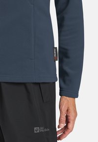 Dunkelblaues Fleece-Jackett mit Polartec-Label an der Seite. Verfügt über einen texturierten Stoff und einen lockeren Schnitt, kombiniert mit schwarzen Hosen.