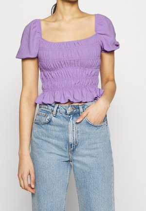 Vrouw die een paarse gerimpelde cropped top met pofmouwen draagt en lichtblauwe hooggetailleerde jeans, met één hand in haar zak.