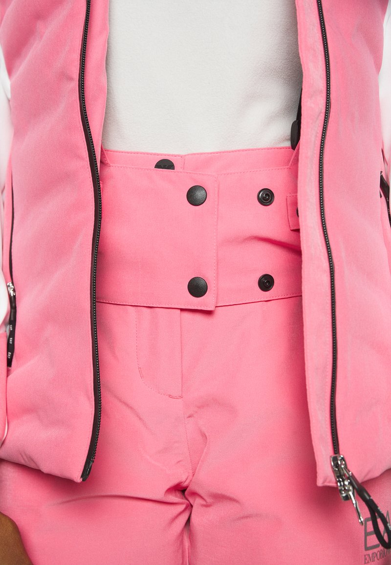 Roze vest met zwarte ritssluitingen over bijpassende hooggetailleerde broek met een knoopsluiting en discrete zakken. Gladde, matte textuur.