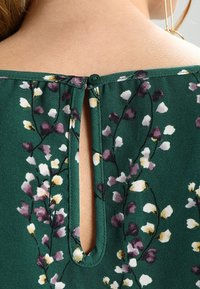 Detalles de una blusa floral verde que presenta una abertura en forma de ojal en la parte posterior, asegurada con un botón verde oscuro, y un tejido suave y texturizado.
