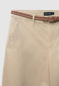 Pantaloni di cotone beige con una texture liscia, dotati di tasche laterali e una cintura intrecciata in pelle marrone. L'etichetta del marchio riporta "CALLIOPE."