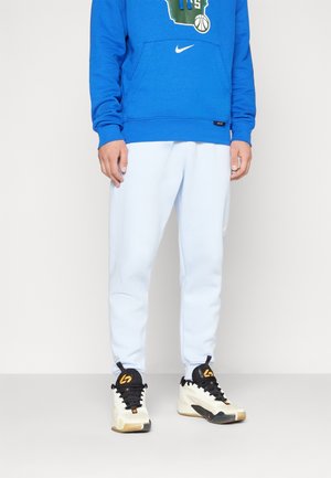 Person iført en blå Nike sweatshirt, lyseblå joggingbukser og hvide, sorte og orange højtop-sneakers stående på en almindelig gulv.