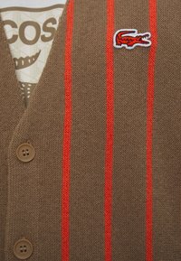 Brązowa swetrowa kamizelka z czerwonymi paskami, z białym haftowanym logo krokodyla i dwoma brązowymi guzikami. Gładka tekstura, wycięcie w kształcie v.