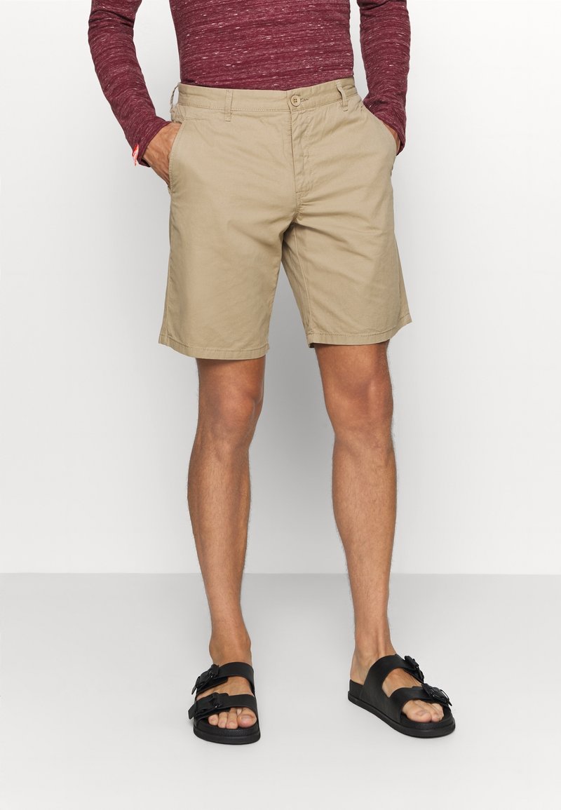 Shorts en coton beige avec une coupe droite, deux poches avant et une fermeture par bouton. Assortis avec des sandales noires à lanières.