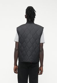 Μαύρο γιλέκο με ραφές και ψηλό γιακά, με υφασμάτινο μοτίβο. Διαγώνια κομμένο στους ώμους και χωρίς μανίκια. Φορεμένο πάνω από γκρι πουκάμισο.