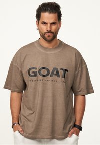 Braunes, oversized T-Shirt mit schwarzem "GOAT"-Text und darunter "GREATEST OF ALL TIME". Weicher Stoff, kurze Ärmel, runder Halsausschnitt. Keine auffälligen Muster.