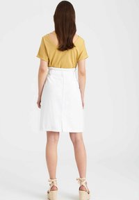 Short-sleeve mustard top with back cutout, paired with a white high-waisted skirt. Model wears tan espadrilles with bow accents.