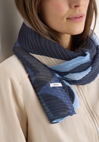 Gros plan sur une femme portant une écharpe rayée bleu marine, bleu clair et beige sur une veste zippée beige.