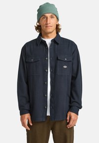 Donker marineblauwe overhemd met knopen, gemaakt van stof en voorzien van twee borstzakken en een logopatch. Gekozen boven een wit T-shirt en olijfgroene broek.
