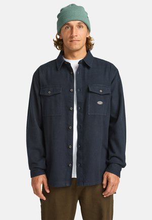 Dunkelblaues Button-up-Shirt aus Stoff mit zwei Brusttaschen und einem Logopatch. Über einem weißen T-Shirt und olivfarbenen Hosen getragen.