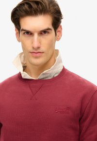 Bordeaux sweatshirts med rund halsudskæring og ribstrikkede manchetter, lagdelt over en skjorte med krave i lys beige, med et broderet logo.
