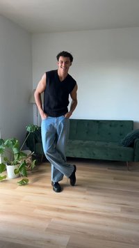 Zwarte mouwloze ribbels top, lichtblauwe baggy jeans en zwarte schoenen. Staand in een lichte kamer met een groene bank en een kamerplant.