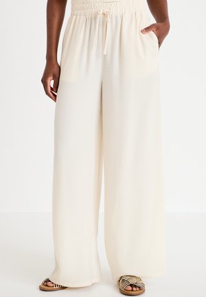 Osoba ubrana w luźne, kremowe spodnie o szerokich nogawkach z wysokim stanem i elastycznym paskiem, oraz beżowe plecione sandały, stojąca z jedną ręką w kieszeni.