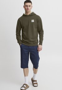 Felpa verde oliva con tasca frontale e toppa con logo, abbinata a pantaloni cargo blu navy. Sandali chiari e calzini bianchi.