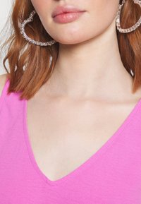 Femme aux lèvres brillantes, cheveux roux, portant de grandes boucles d'oreilles en forme de cœur en cristal et un haut rose à col en V.