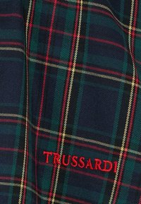 Tessuto tessuto con un motivo scozzese blu scuro e verde, con accenti rossi e gialli. "TRUSSARDI" ricamato in rosso.