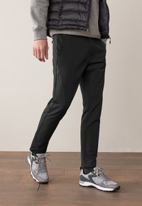 Pantalon noir fuselé avec des fermetures éclair sur les poches, associé à des chaussures de sport grises avec des accents noirs et des lacets gris. Fond en parquet.