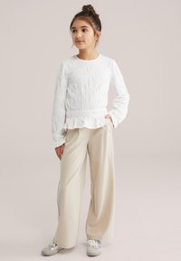 Haut blanc à manches longues texturé avec un ourlet à volants, associé à un pantalon large rayé beige et des baskets argentées. Tenue simple et décontractée.
