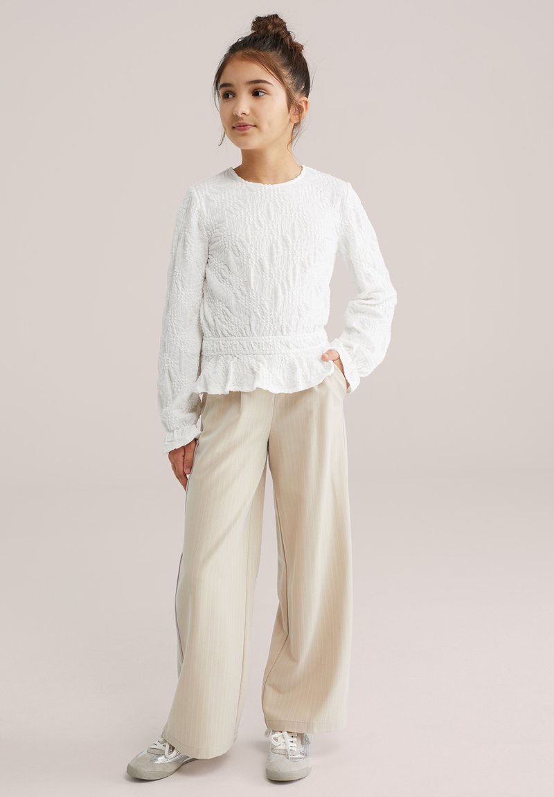 Haut blanc à manches longues texturé avec un ourlet à volants, associé à un pantalon large rayé beige et des baskets argentées. Tenue simple et décontractée.