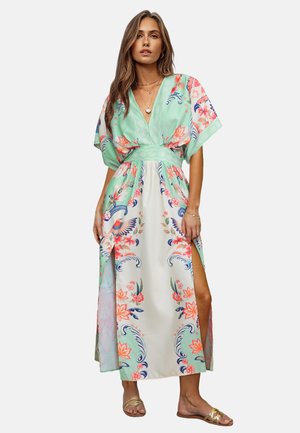 Mujer con un vestido largo floral con patrones en verde, blanco y rosa, que presenta un escote en V profundo, mangas anchas y aberturas en la parte delantera de las piernas.