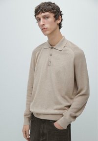 Polo beige in maglia con colletto, abbottonatura a tre bottoni e maniche raglan. Texture morbida e bordi a costine, indossata con jeans scuri.