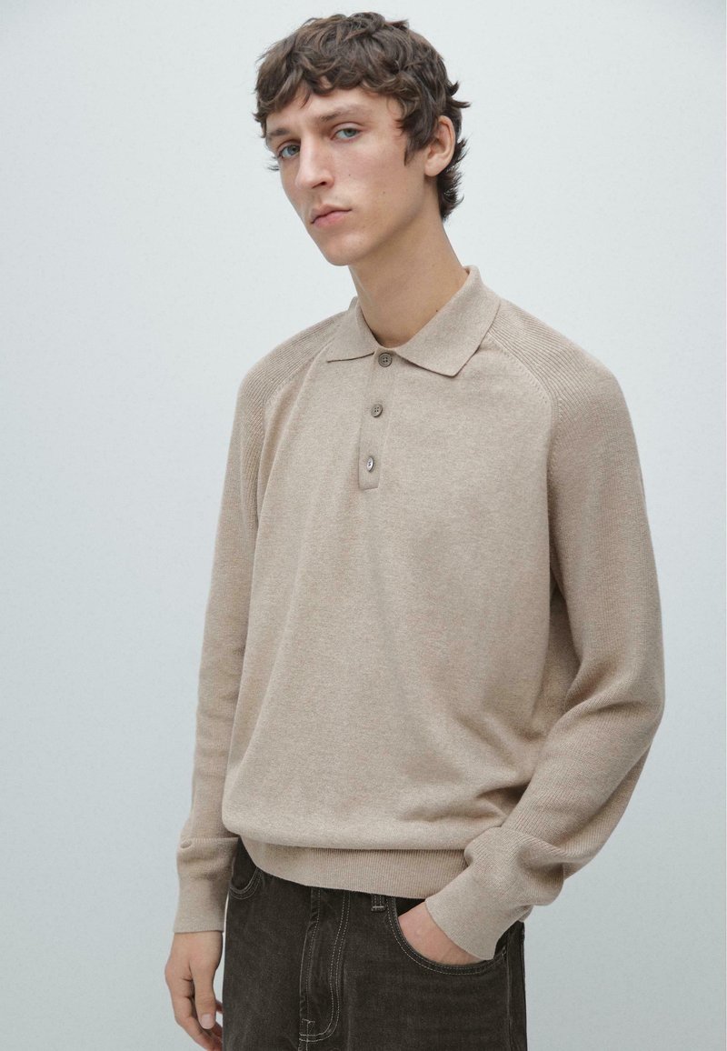 Polo beige in maglia con colletto, abbottonatura a tre bottoni e maniche raglan. Texture morbida e bordi a costine, indossata con jeans scuri.