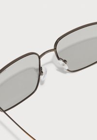 Ochelari de soare metalici cu lentile dreptunghiulare de un gri închis. Rama este de culoare bronz, cu o textură subtilă, având pernuțe nazale transparente și tije subțiri.
